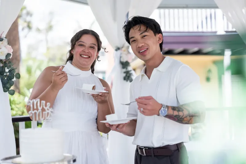 The two smile at each other, each holding a plate of cake and a champagne glass in the bright venue space.