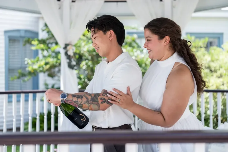 Two people in white attire hold a champagne bottle and smile on the covered porch.