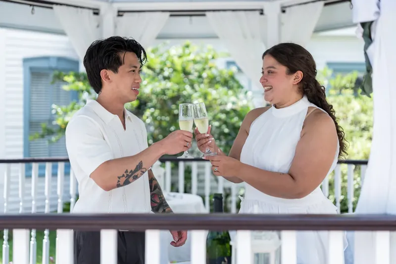 Blossom and Chris clink champagne glasses together, both grinning at the railing overlooking the grounds.