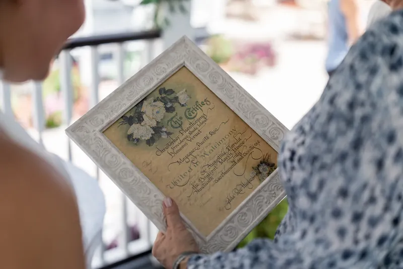 Someone in a white dress holds a framed marriage license while standing on a sunny porch.