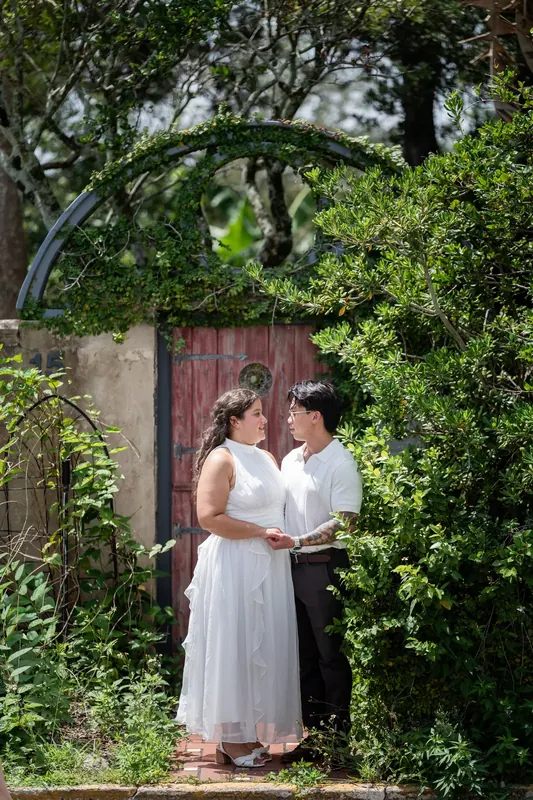 Two people in white stand facing each other beneath a green ivy-covered archway with a burgundy door.