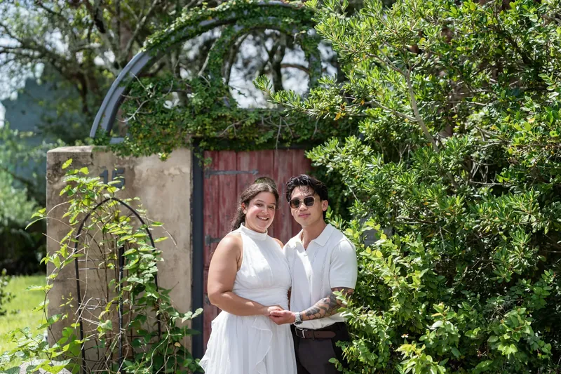 The couple stands arm-in-arm under lush vines, smiling at the camera near a weathered red entrance.