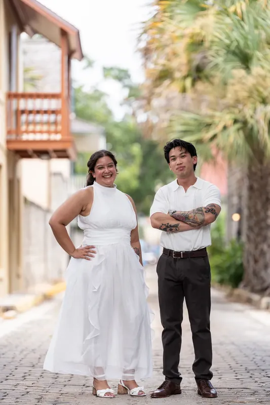 The two pause on a tree-lined cobblestone path, one with hand on hip, sharing a relaxed moment downtown.