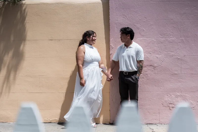 One person in white linen and another in a white t-shirt stand close against a cream and mauve wall.