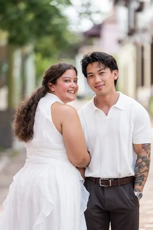 Two people stand shoulder-to-shoulder on a quiet street, smiling at the camera in soft afternoon light.