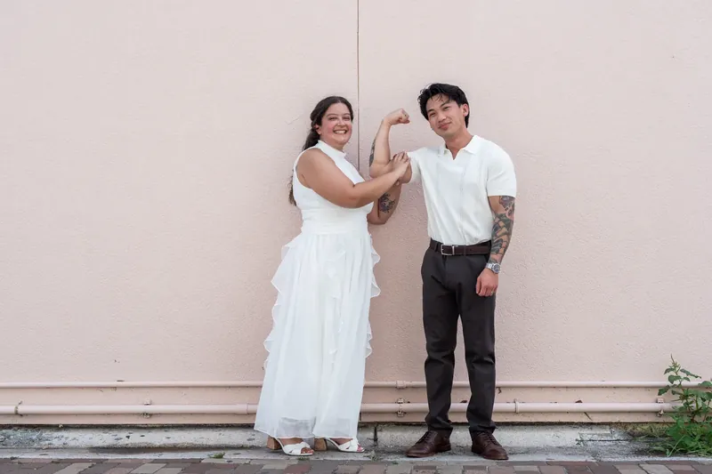 One partner gently adjusts the other's white gown against a pale concrete wall, a quiet moment of care between the two.