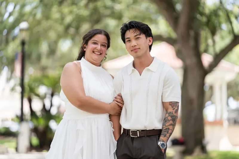 Blossom and Chris walk arm-in-arm smiling under the oak canopy, Chris showing a visible tattoo on his arm.