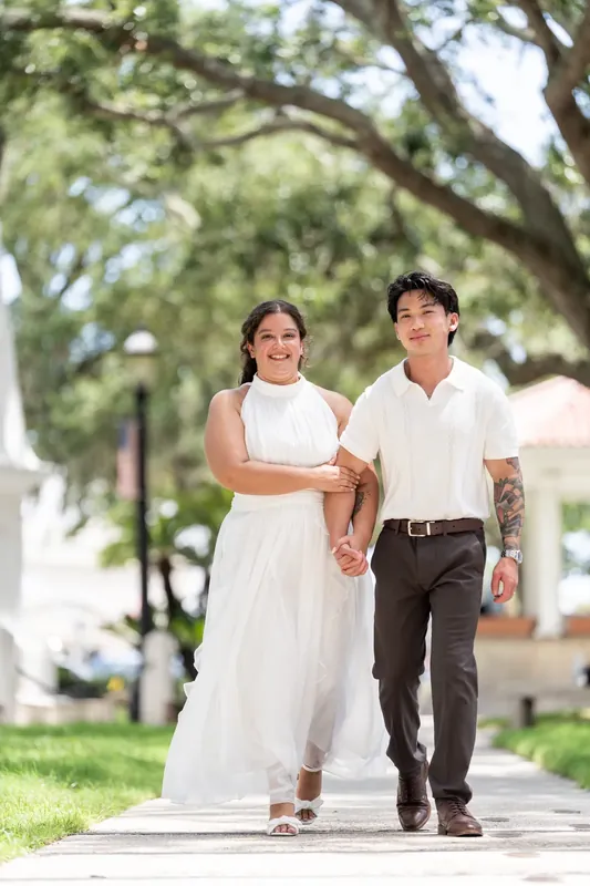 Two people walk hand-in-hand down a tree-lined brick pathway beneath mature oaks in downtown St. Augustine.