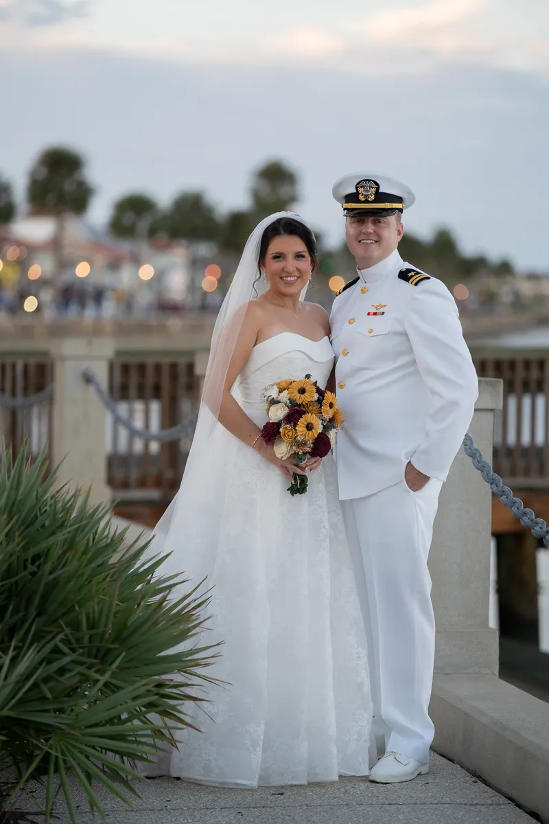 Brianna holds a yellow bouquet while George stands beside her in his white dress uniform on the bayfront promenade