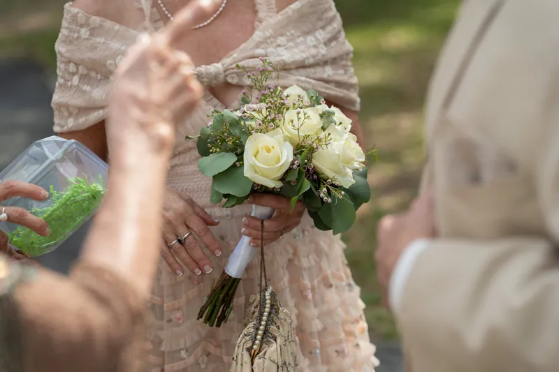 Bridgid holds a white rose bouquet with trailing ribbon while wearing a lace dress with a bow detail at the shrine.