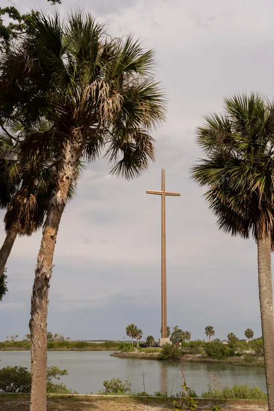 Tall wooden cross stands between palm trees overlooking water under a cloudy sky at the mission grounds.