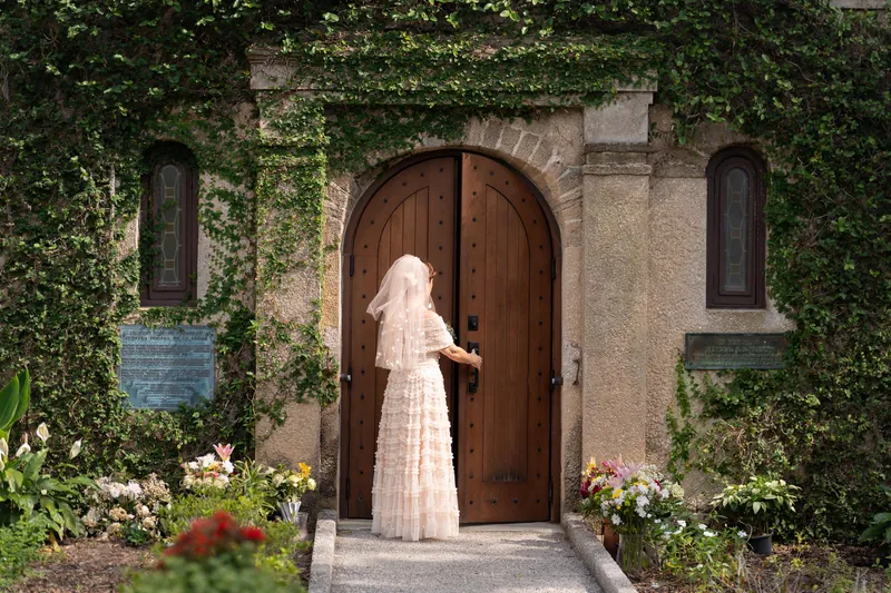 Bridgid in her lace wedding dress and veil stands at the mission's wooden entrance door, flanked by flowering arrangements.