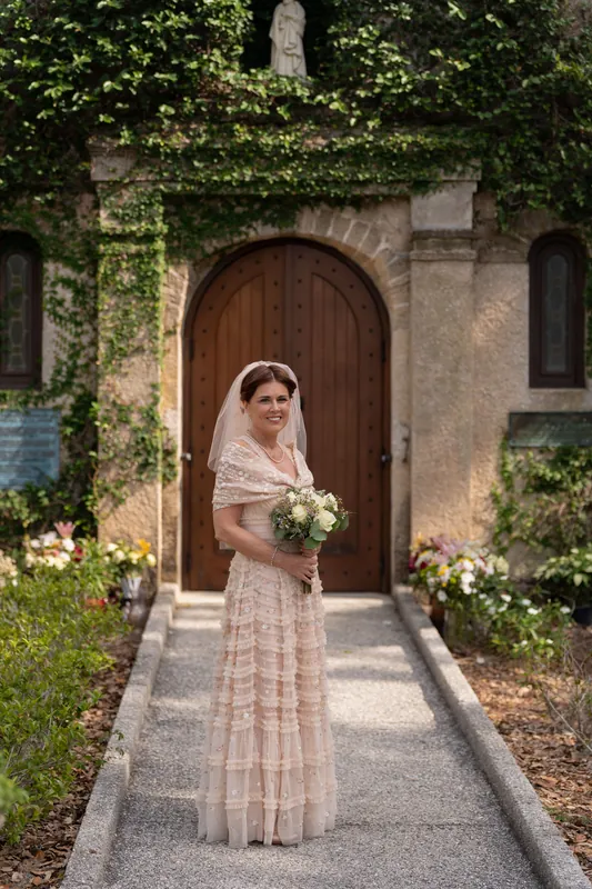 Bridgid stands on a shaded pathway in front of climbing ivy and an arched doorway, holding cream and green flowers.