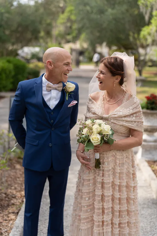 Dan and Bridgid face each other smiling on a gravel path surrounded by trees, holding white and green blooms.
