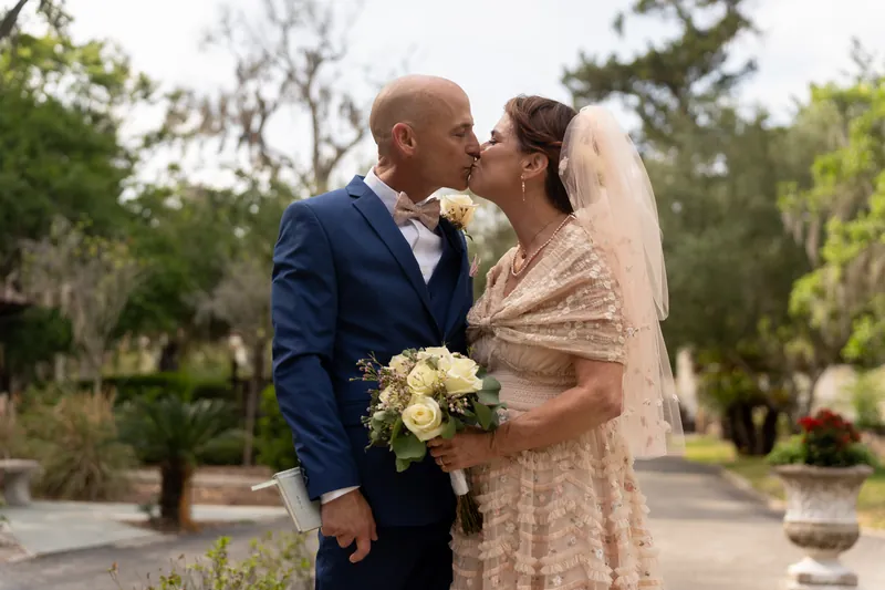 Dan leans in to kiss Bridgid as she holds her bouquet, both dressed formally beneath the canopy of ancient oaks.