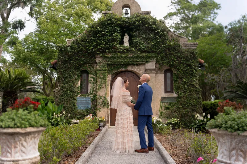 Bridgid and Dan face each other on a brick path flanked by flowering gardens and an ivy-draped stone chapel.