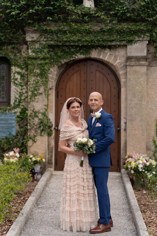 Bridgid holds a white bouquet while Dan embraces her on the brick walkway, ivy-covered archway framing them behind.