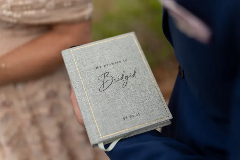 A cream-colored vow booklet printed with "Bridgid" sits open in Dan's hands as he holds it during the ceremony.