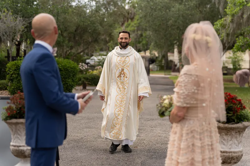 A priest in white vestments conducts the ceremony as Dan and Bridgid stand before him among flowering planters.