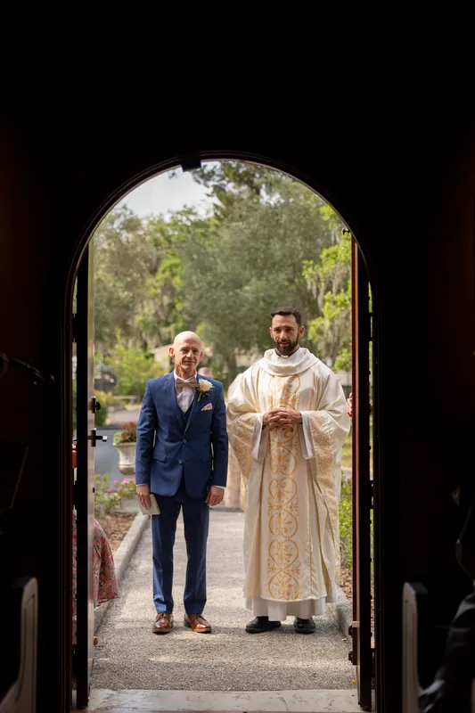 Dan emerges through the wooden arched doorway toward the priest waiting on the sunlit grounds outside.