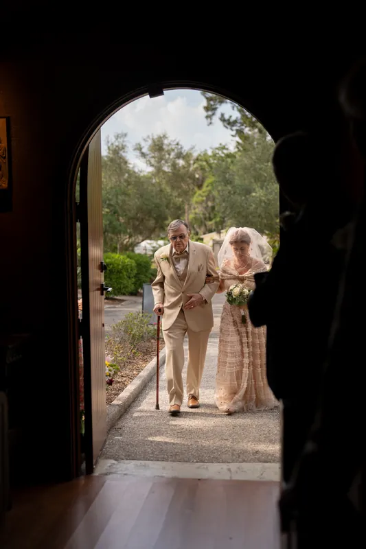 Framed by a wooden doorway, Dan and Bridgid walk together down a garden path lined with greenery at the mission shrine.