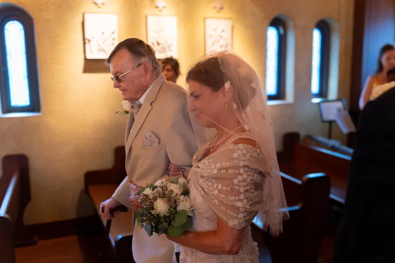 Inside the candlelit chapel, Bridgid holds a bouquet while Dan stands beside her in a tan suit during the ceremony.