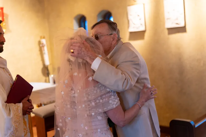 A man in a tan suit embraces a woman wearing a lace veil and beaded gown within the warmly lit stone chapel.