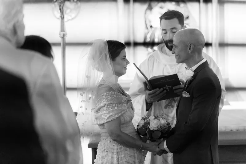 Black and white photograph of Bridgid and Dan reading from a book during their ceremony, with a priest presiding nearby.