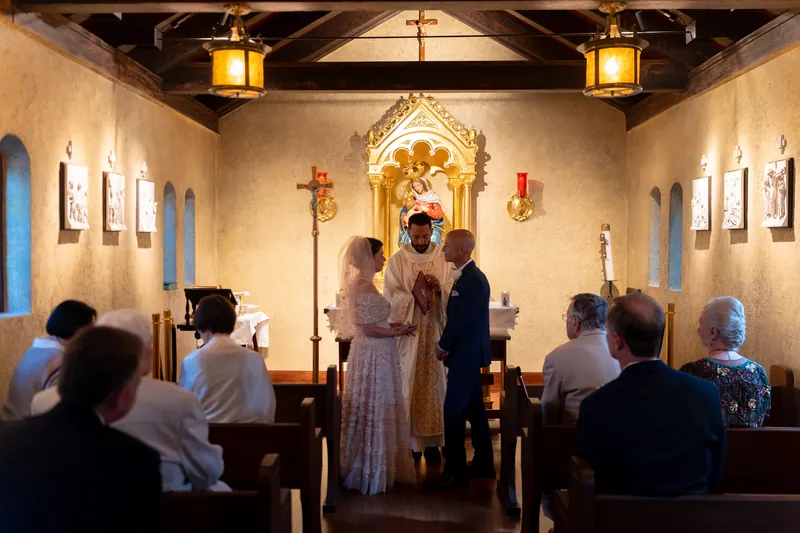 Bridgid in a cream lace dress exchanges vows with Dan in a navy suit as guests look on in the candlelit chapel.