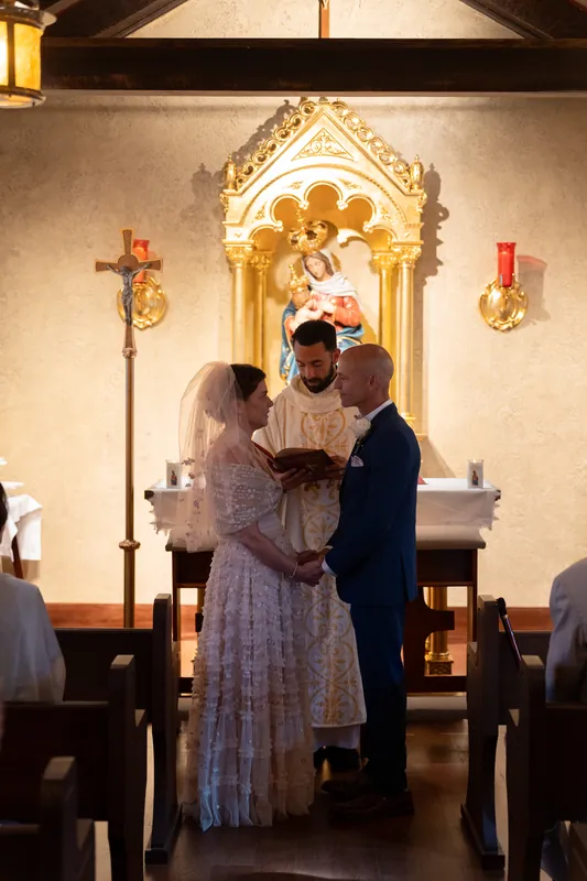 Dan slips a ring onto Bridgid's finger while she holds her veil, a priest officiating between them.