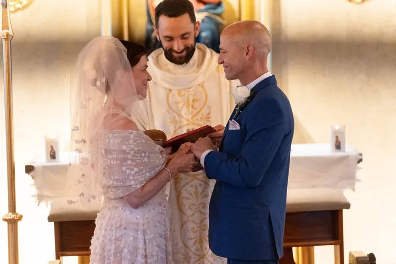 The couple clasps hands during their vow exchange, with the officiant holding their joined hands as witnesses sit nearby.