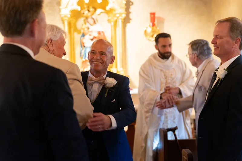 Dan converses with guests in the Mission's golden-lit chapel, wearing a dark suit with a boutonniere.