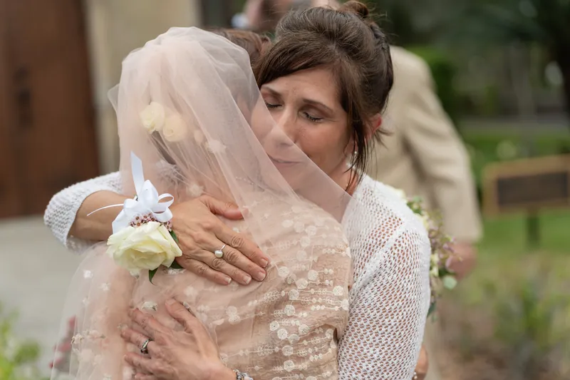 Bridgid smiles beneath her veil while hugging someone outside the Mission, holding a white bouquet.