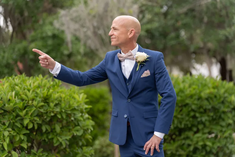 Dan gestures expressively in the Mission's grounds, dressed in a navy suit with green foliage behind him.