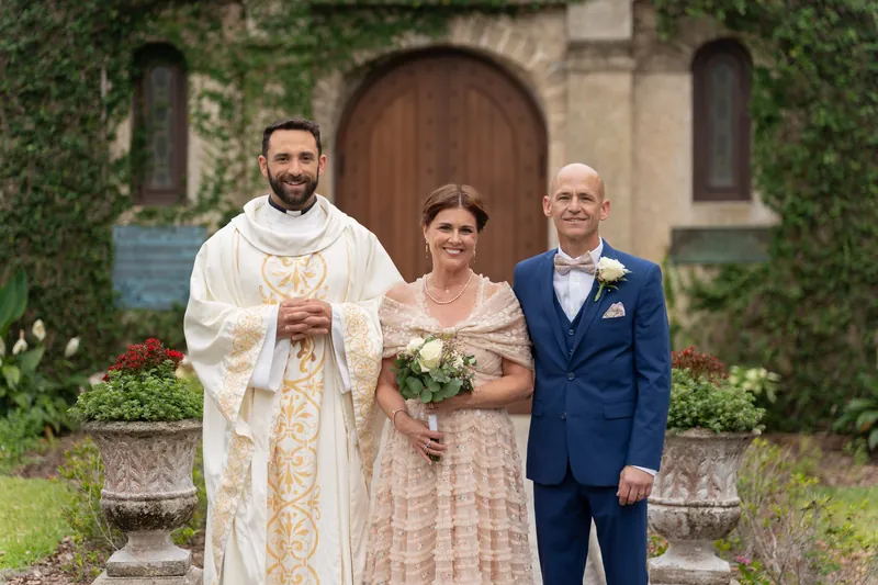 A priest in cream vestments stands with Bridgid and Dan before the ivy-covered shrine entrance.