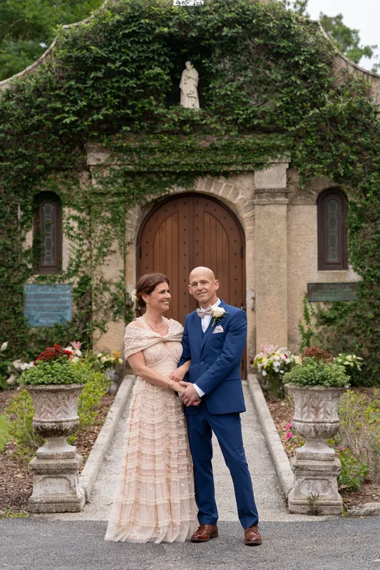 Dan in navy and Bridgid in her pleated pale pink gown hold hands facing the wooden arched doorway framed by climbing ivy.