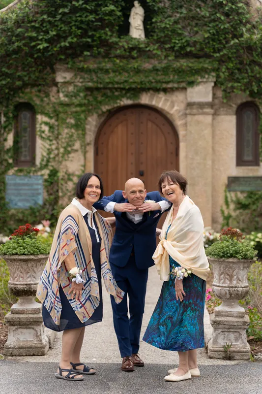 Two guests flank Dan in a navy suit outside the ivy-covered shrine, adjusting his jacket with warm smiles.