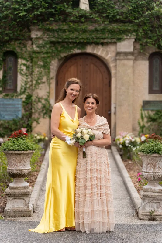 Bridgid in a yellow gown and a guest in peach stand together holding a white bouquet before the stone chapel.