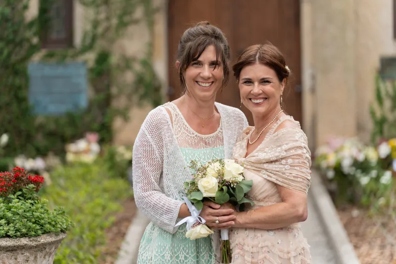 Bridgid and a companion hold a cream-and-green bouquet, both smiling in the courtyard's garden setting.