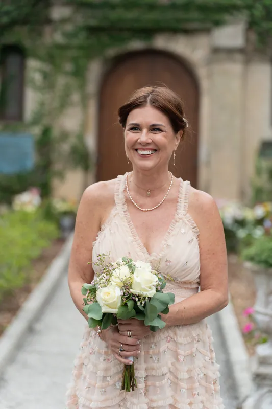 Bridgid in a blush pleated gown clutches her bouquet and grins at the camera, ivy-draped walls behind her.