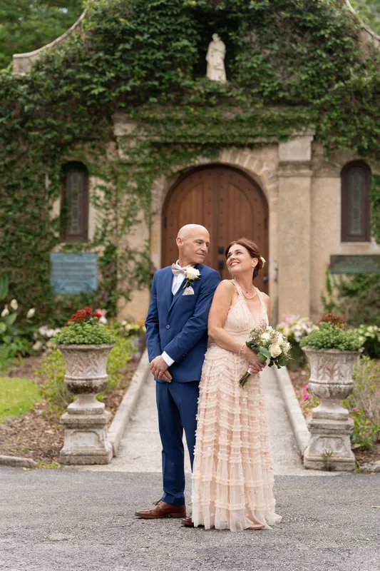 Dan and Bridgid walk arm-in-arm down a stone path with ivy-covered walls and a chapel entrance behind them.