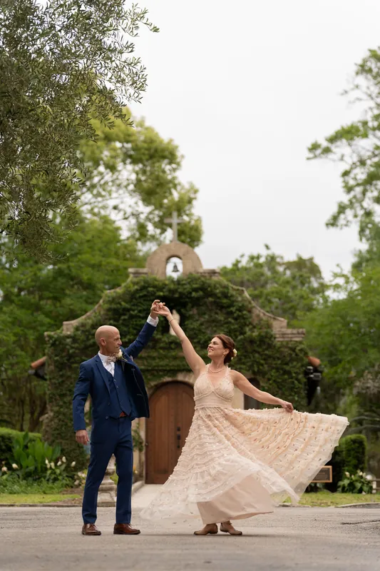 Bridgid twirls her flowing skirt while Dan watches, the historic shrine's bell tower visible behind them among mature trees.