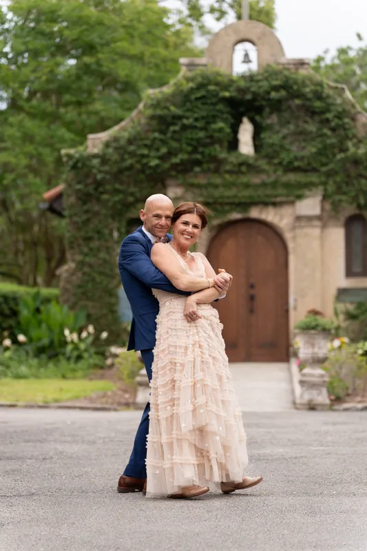 Dan embraces Bridgid from behind as they stand before the ivy-draped Mission building, both smiling at the camera.