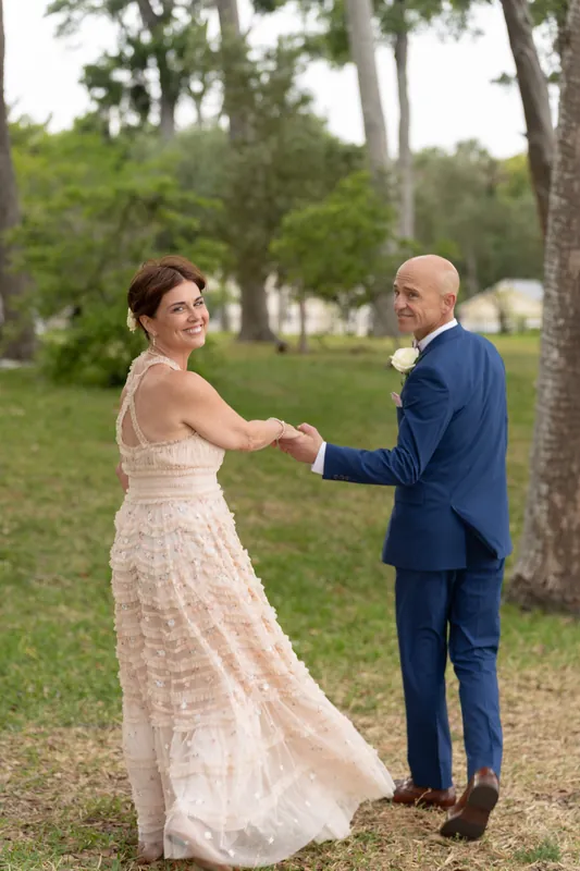 Bridgid in a cream tiered gown and Dan in navy hold hands beneath tall oaks at Mission Nombre de Dios.