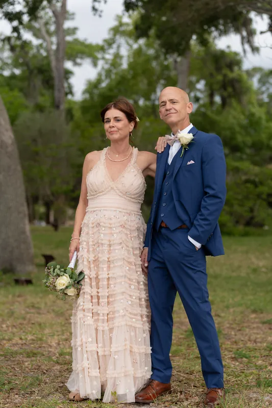 Bridgid and Dan stand side by side on the grounds, she in cream lace and he in navy, holding a white bouquet.