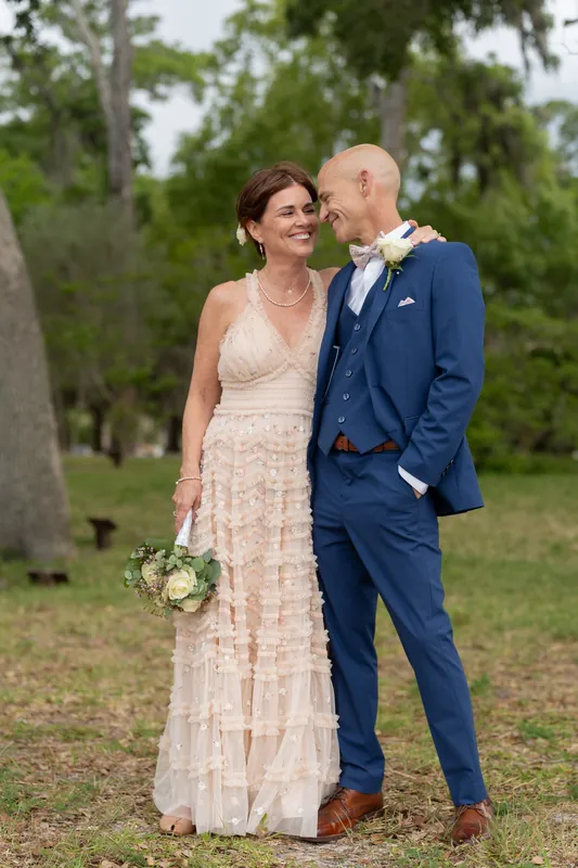 The couple walks together beneath the oaks, Bridgid in her layered cream dress and Dan in navy, carrying their flowers.
