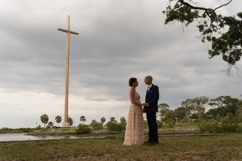 Bridgid in a cream checkered dress and Dan in navy stand beneath a tall wooden cross on the shrine grounds.