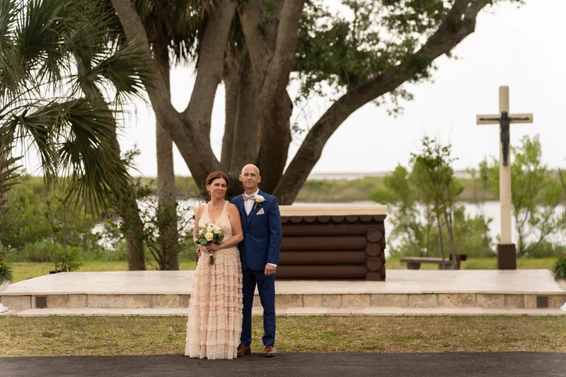 Bridgid and Dan pose together under a sprawling oak tree with a wooden bench and cross behind them.