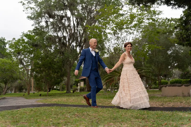 Bridgid and Dan hold hands walking across the manicured grounds, mature trees creating a natural canopy overhead.