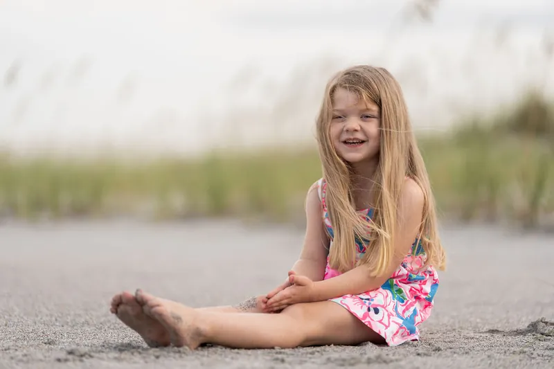 Girl in floral dress sits on the sand, grinning at Vilano Beach.